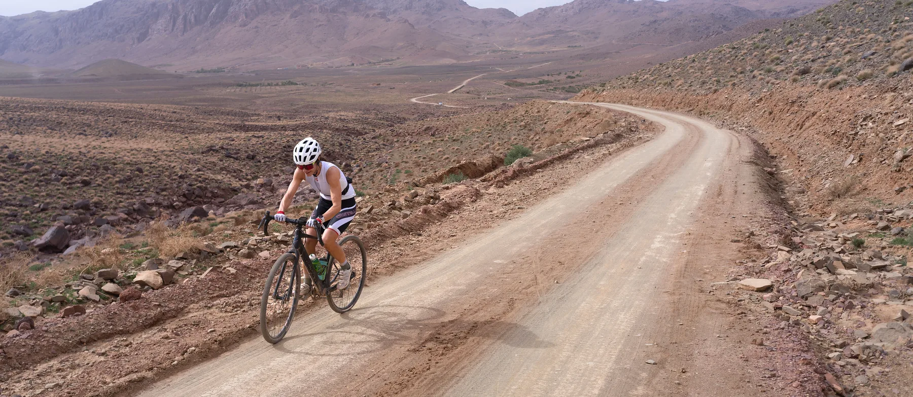 A lone cyclist on a wide dirt road crossing a bare valley, the road running on to the horizon