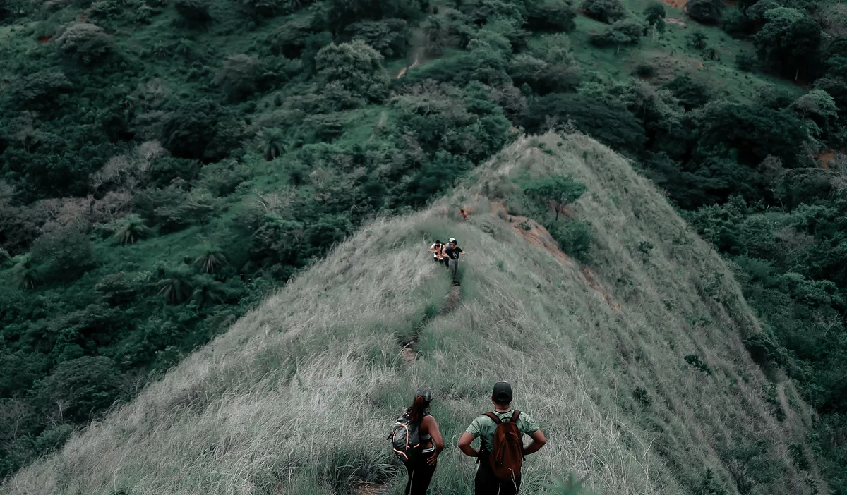 Two hikers on a narrow grass ridge, seen from above, with green slopes falling away on both sides