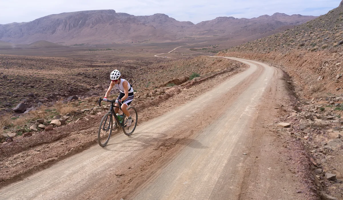 Gravel cyclist on a wide dirt road crossing a bare valley under a line of mountains